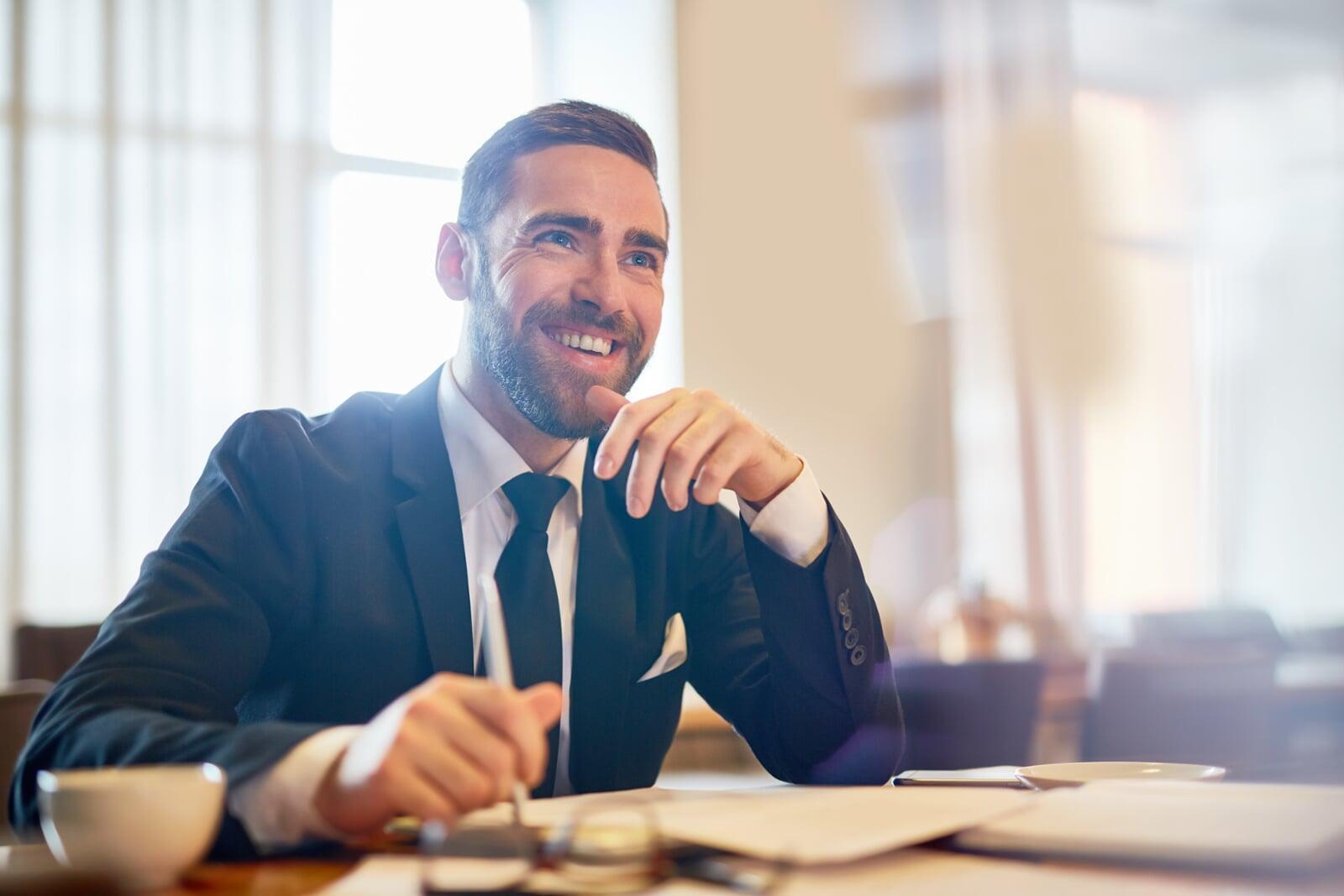 Man sitting in a restaurant in a suit, looking happy with papers on the table in front of him. Owning his personal brand.