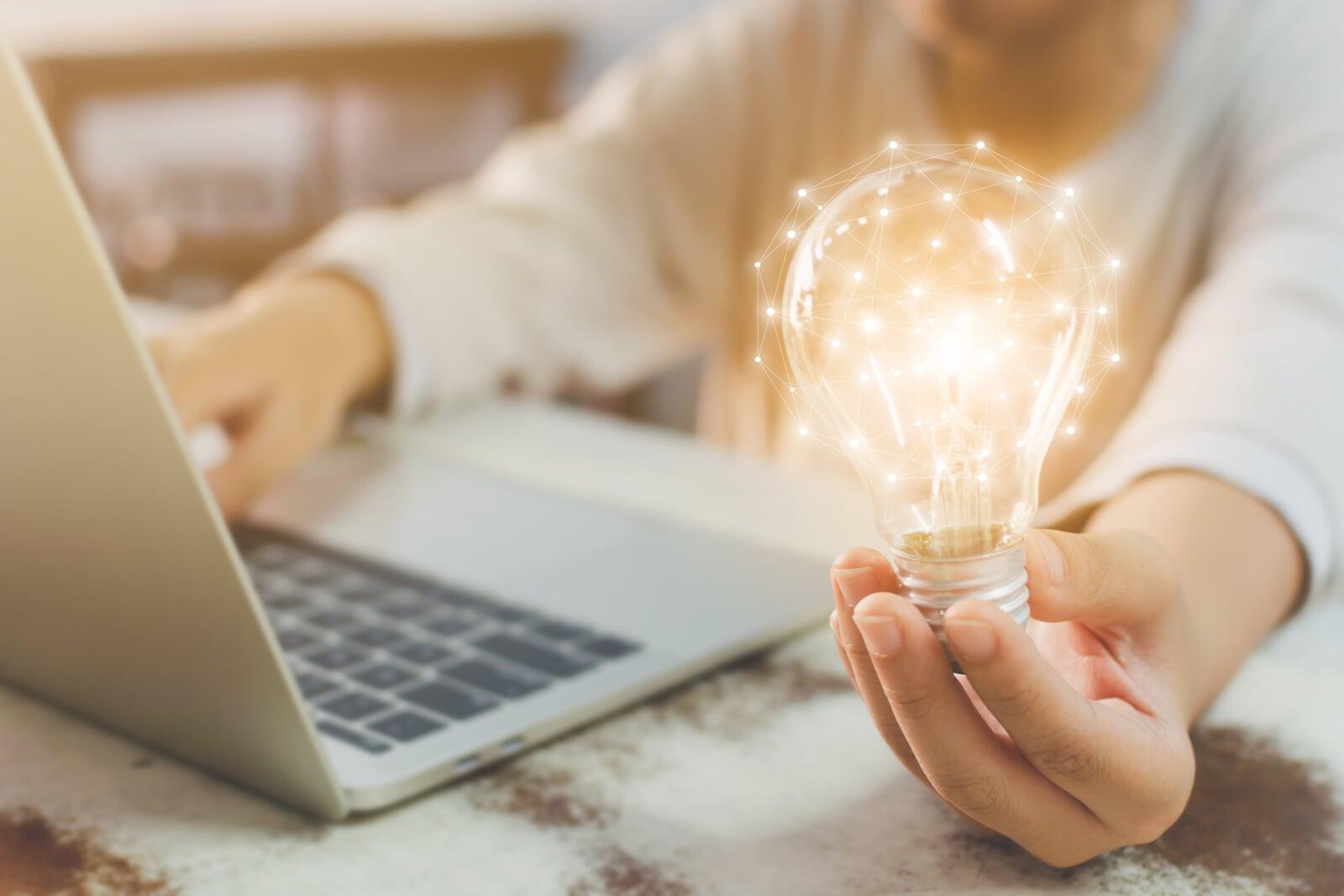 Person sitting at a desk with a laptop, with a light bulb image in hand, highlighting a breakthrough.