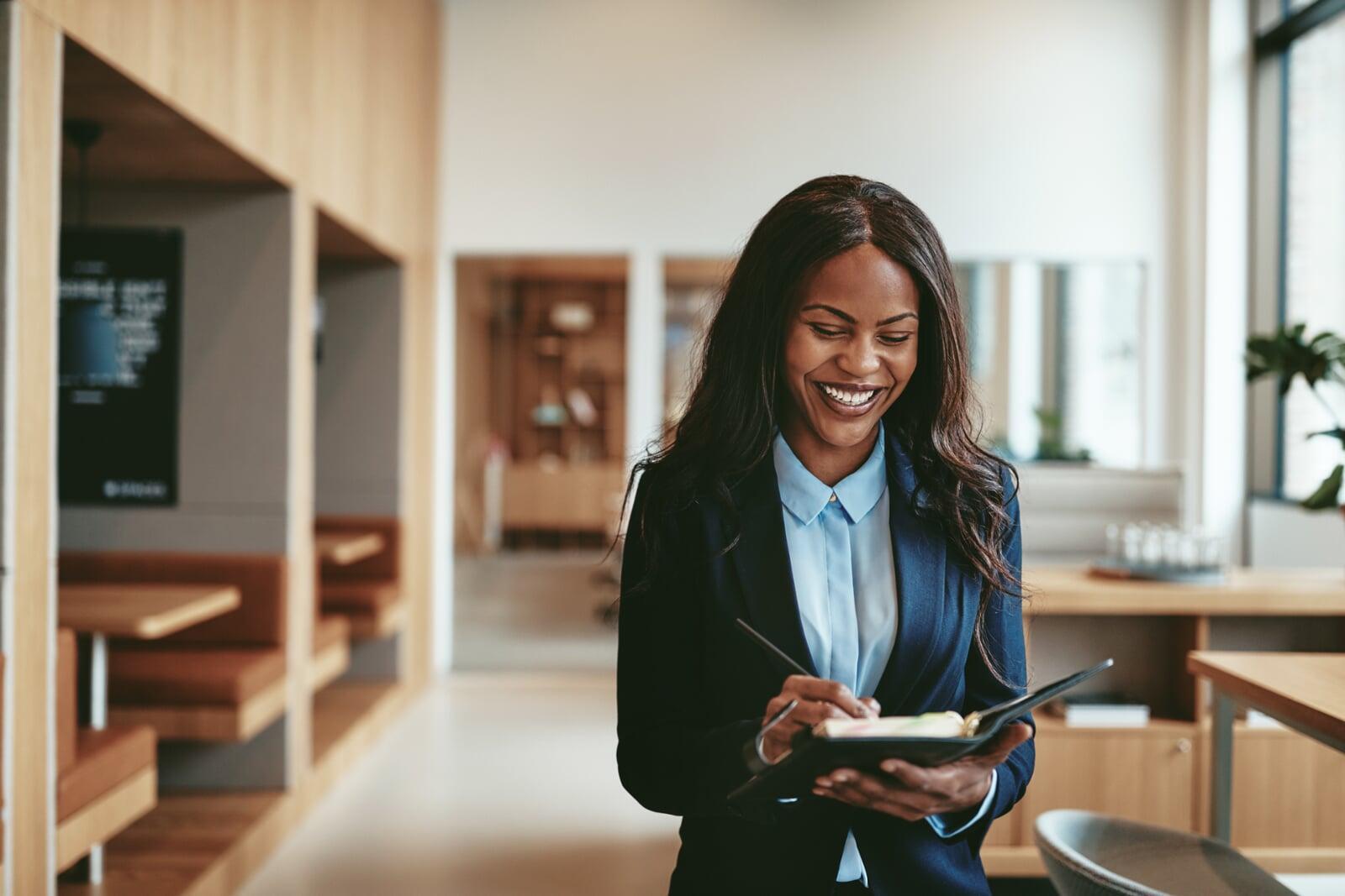 Woman standing in a cafeteria type setting, with a notebook looking happy and optimistic about the future.